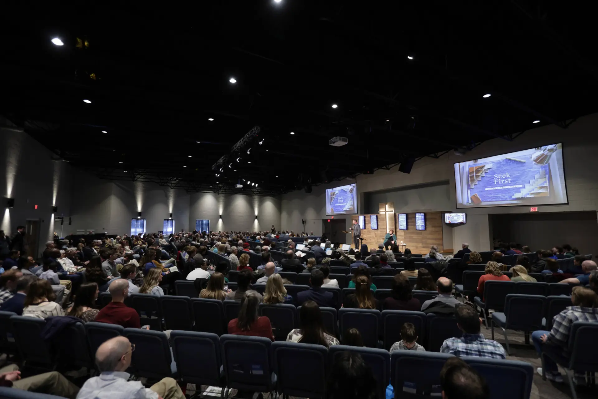 A large group of people sits in rows of chairs inside a church auditorium, watching a speaker on stage with presentation slides projected on screens.