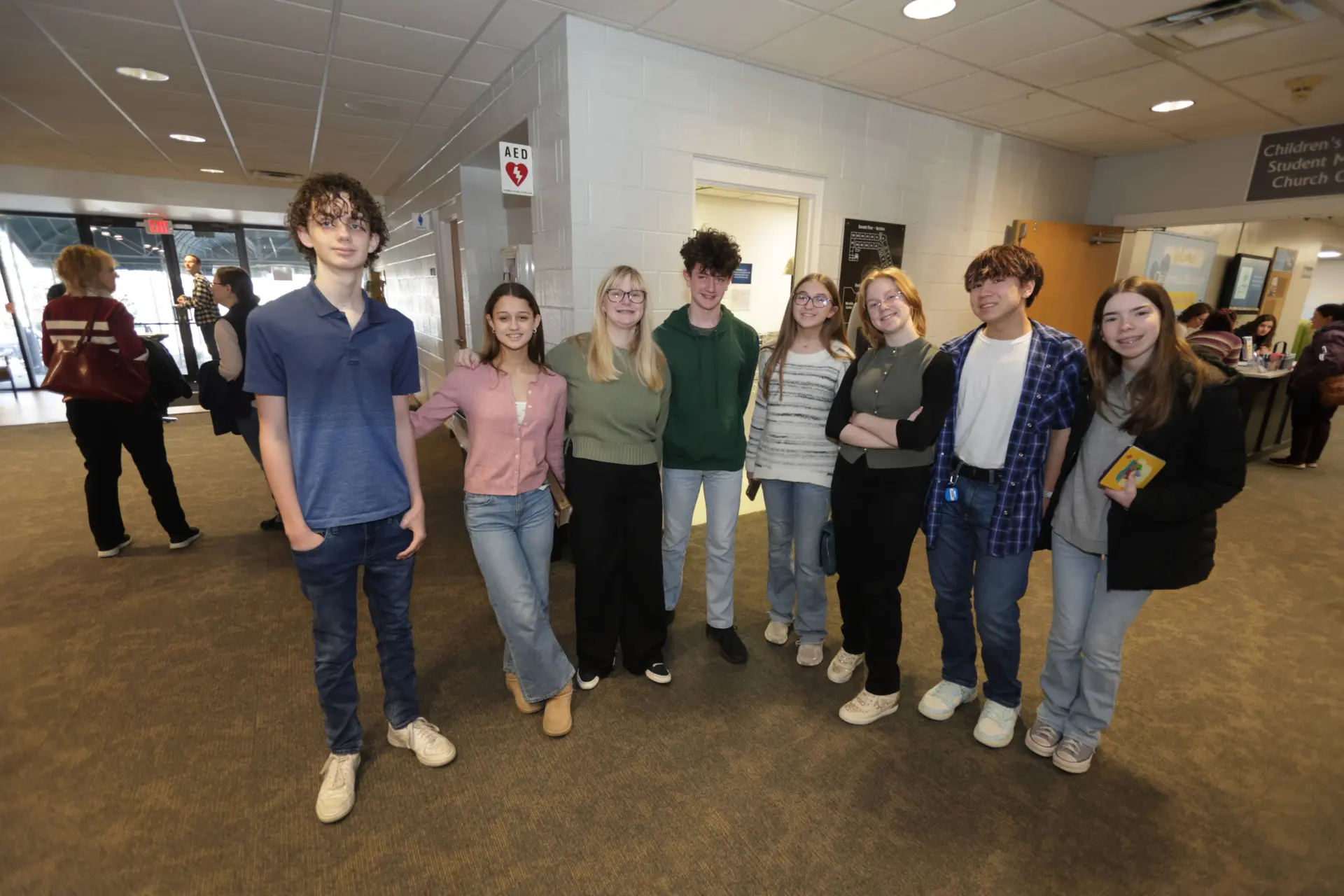 A group of eight teenagers stands together indoors, posing and smiling for the camera in a public hallway with other people in the background.
