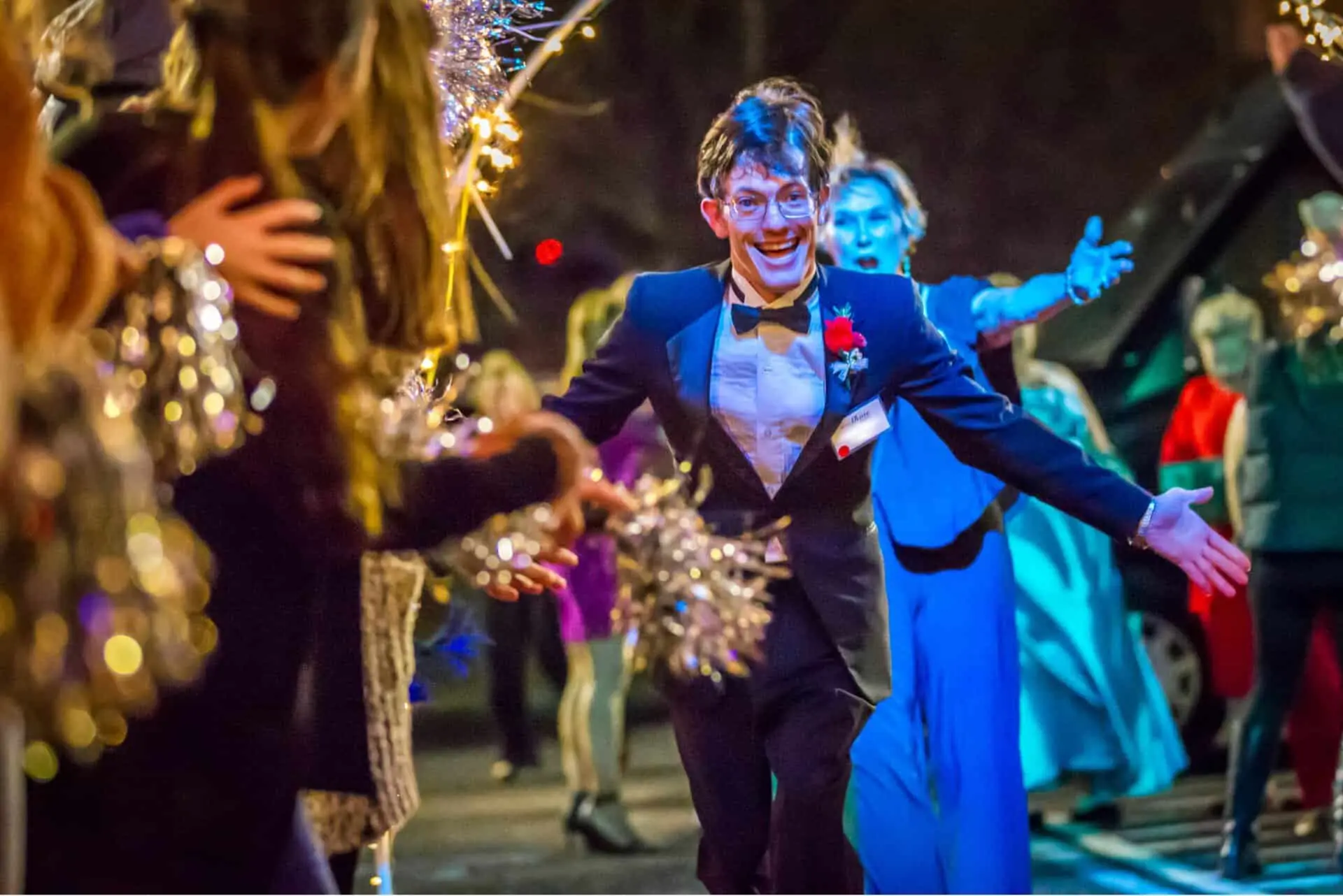 A young person in formal wear runs down an outdoor path lined with people cheering and holding shiny pom-poms, at night.